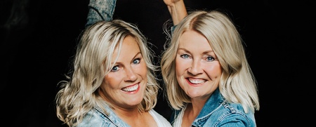 Två glada kvinnor i ljusa kläder och jeansjackor poserar framför en mörk grottöppning.