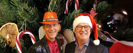 Två män står framför en julgran, båda bär julhattar och ler mot kameran.
