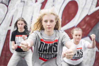 Tre unga personer dansar energiskt framför en graffitivägg, med fokus på en tjej i grå t-shirt.