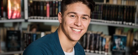 Ung man sitter leende på en blå stol i ett bibliotek med bokhyllor i bakgrunden.
