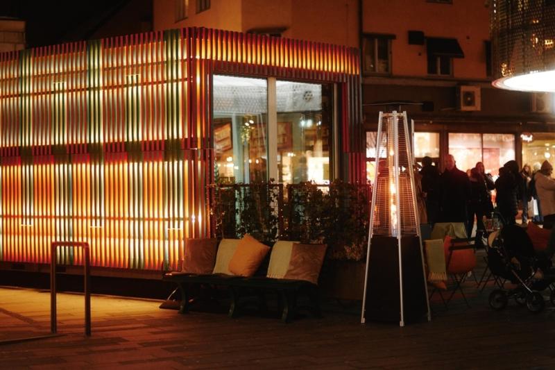 Uteservering med färgglada ljus, sittplatser och värmare på kvällen, folk i bakgrunden.