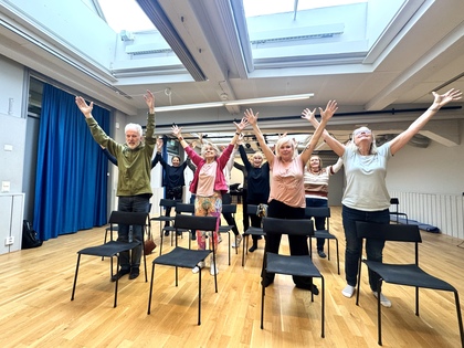 En grupp äldre personer står bakom stolar i ett ljust rum och sträcker armarna uppåt, som i en gruppövning.