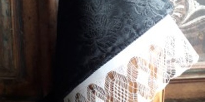 Svart spetsduk med blommönster och vit spetskant, placerad på en träyta framför en dekorerad bakgrund.