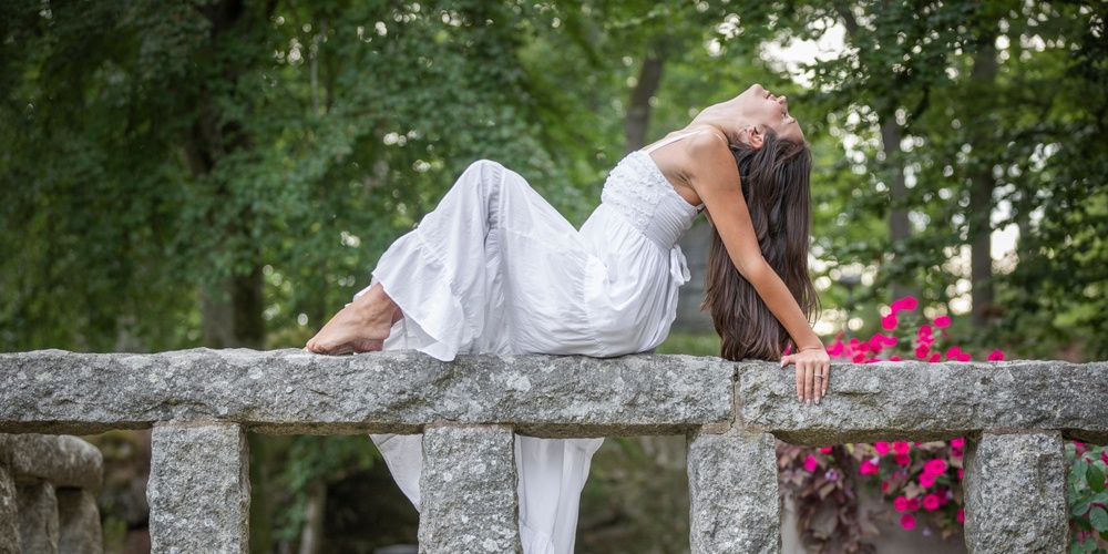 Kvinna i vit klänning poserar liggande på ett stenräcke i en grönskande park.