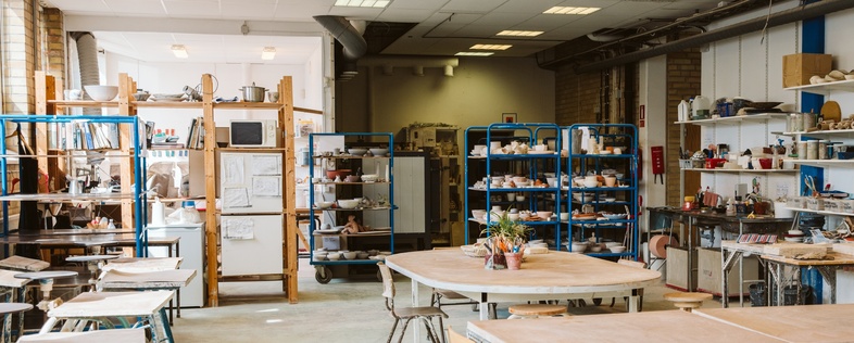 Ljust keramikverkstad med arbetsbord, hyllor fyllda med material och verktyg, inga människor syns.