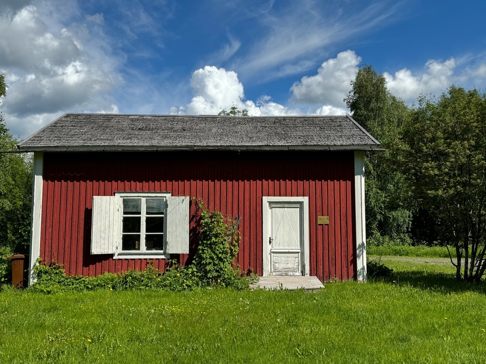 Röd liten stuga med vita knutar, vita fönsterluckor och dörr, omgiven av grön gräsmatta och träd under blå himmel.