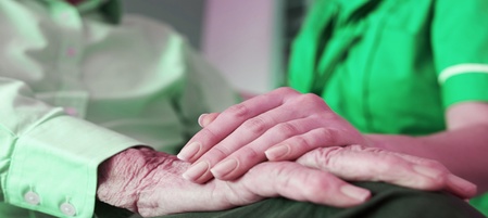 En yngre person håller en äldre persons hand i en stödjande gest. Båda bär gröna kläder.