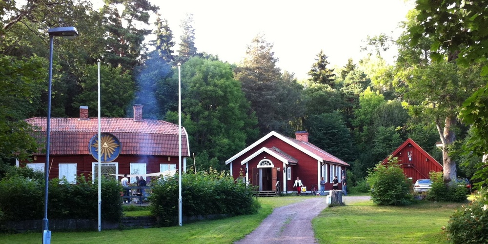 En grusväg leder till tre röda hus omgivna av grönska och träd, med flaggstänger och en grillplats.