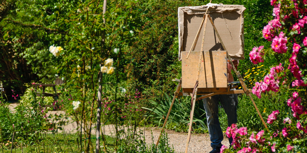 En person målar utomhus i en blomstrande trädgård, duken täcker ansiktet.