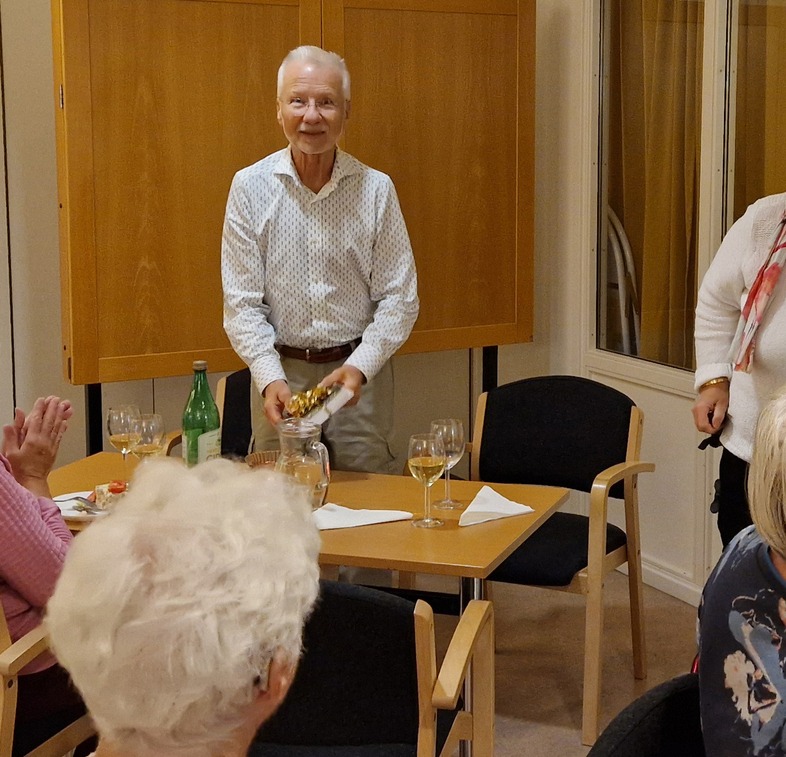 En grupp äldre personer sitter runt ett bord med dryck och mat i ett mötesrum.