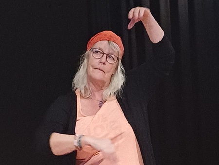 Kvinna med glasögon och röd sjal på huvudet sitter på knä och gestikulerar på en scen med tända ljus och tyg framför sig.