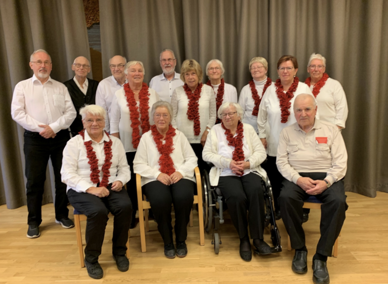 Gruppfoto av äldre personer i vita skjortor och röda halsband, sittande och stående framför en grå gardin.