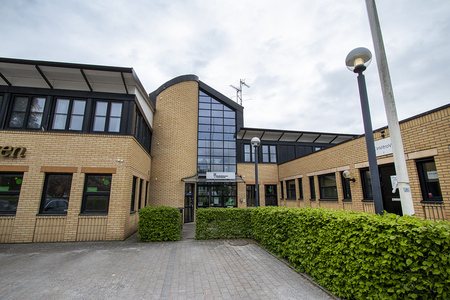 Entré till en modern byggnad med ljusgul tegelfasad, stora fönster och glasparti i mitten.