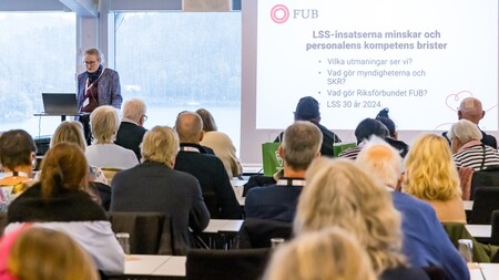 Föreläsare talar inför publik i konferensrum med presentation om LSS på stor skärm.