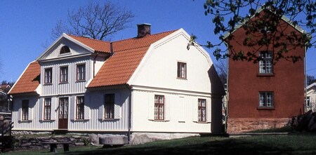 Två traditionella svenska hus, ett vitt trähus och ett rött stenhus, står bredvid varandra under klarblå himmel.