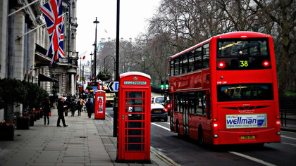 En röd dubbeldäckarbuss och en röd telefonkiosk på en gata i London med brittiska flaggor och människor på trottoaren.
