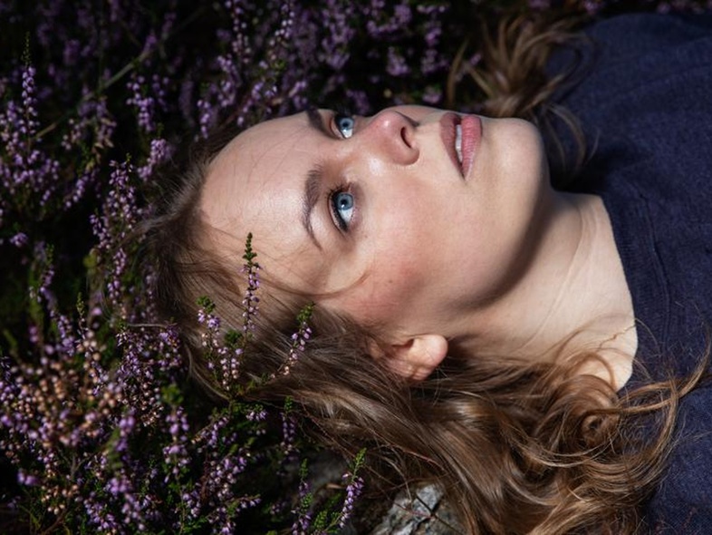 Kvinna med blå ögon ligger på rygg bland lila blommor och tittar uppåt.