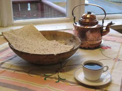 Ett bord med en kopp kaffe, en koppartekanna och ett träfat med knäckebröd framför ett fönster.