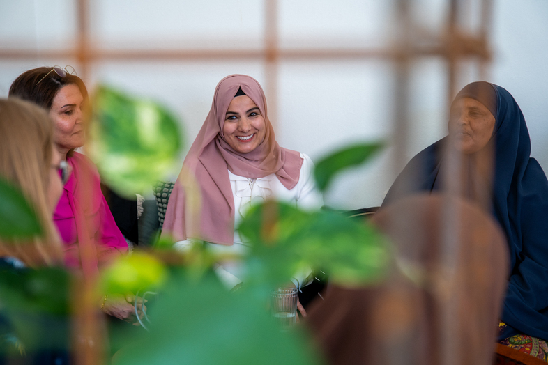 Fyra kvinnor sitter tillsammans och samtalar, en av dem bär rosa hijab och ler. Växter syns i förgrunden.