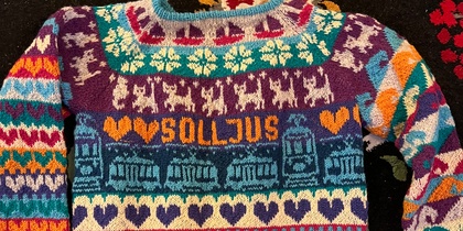 Färgglad stickad tröja med mönster, text och symboler på en blommig matta.