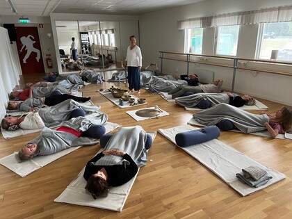 En grupp människor ligger på yogamattor med filtar i en ljus sal, en instruktör står i mitten.