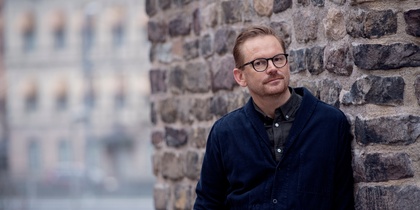 Man med glasögon lutar sig mot en stenmur utomhus, bakgrund med suddiga byggnader.