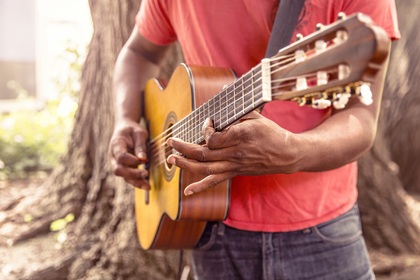 En person spelar akustisk gitarr utomhus, nära ett träd, iförd röd tröja och jeans.