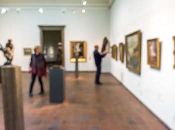 Suddig bild av personer som tittar på konstverk i ett galleri med tavlor och skulpturer.
