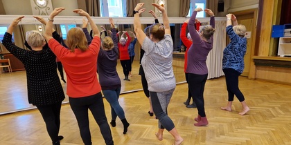 En grupp äldre kvinnor övar balett tillsammans i en spegelsal, alla står med armarna uppsträckta.