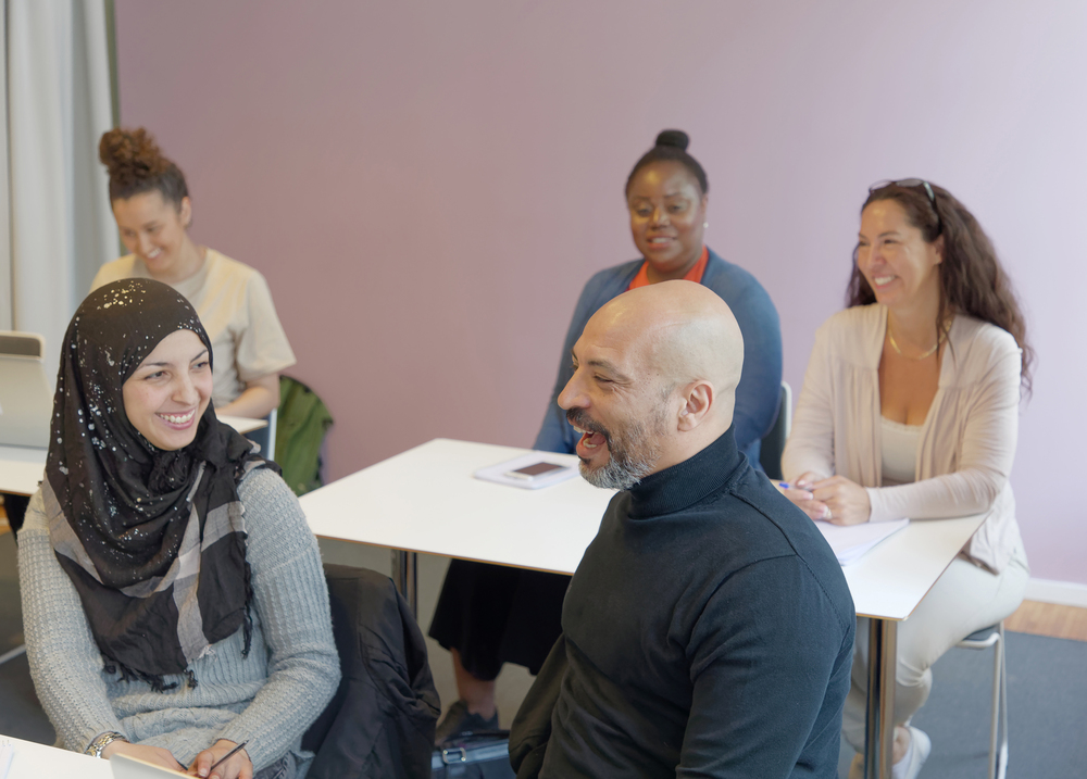 Fyra vuxna sitter vid bord i ett klassrum och samtalar, alla ser glada ut.