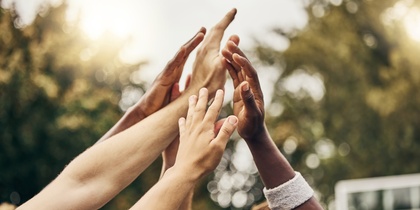 Flera händer från olika personer möts i en high five utomhus, med träd i bakgrunden.