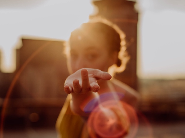 Person står utomhus i motljus med utsträckt hand mot kameran, bakgrunden är suddig.