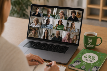 Person deltar i videomöte på laptop, antecknar i block, grön kopp och bok på bordet.