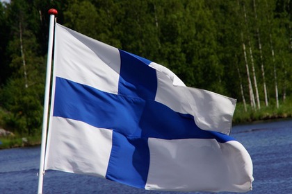 Finlands flagga vajar utomhus vid en sjö med skog i bakgrunden.