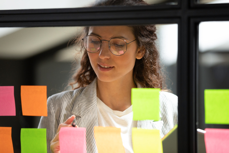 Kvinna med glasögon skriver på färgglada post-it-lappar på en glasvägg på ett kontor.