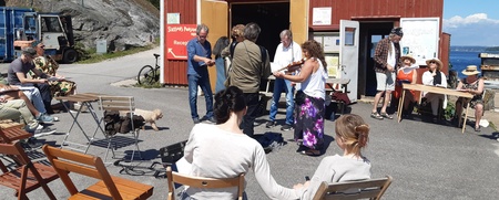 En grupp människor samlade utomhus vid en röd byggnad, några sitter på stolar och andra står och pratar.