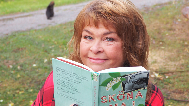 Kvinna med ljust hår sitter utomhus och ler medan hon håller upp en bok med titeln 'Sköna Maj'.