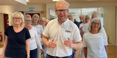 En grupp äldre personer och en yngre man står tillsammans inomhus, leende och avslappnade.