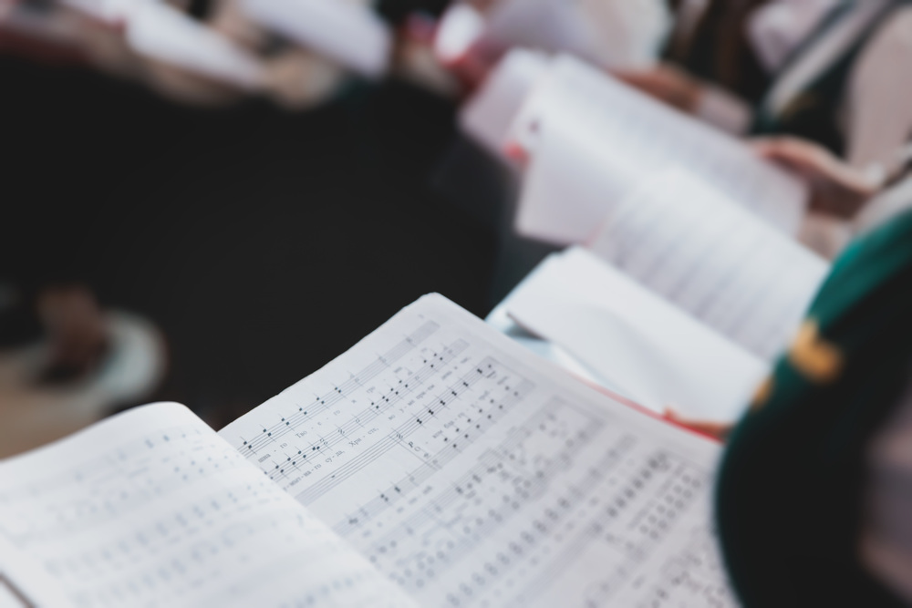 Närbild på ett notblad i en kör, med suddiga personer som håller sånghäften i bakgrunden.