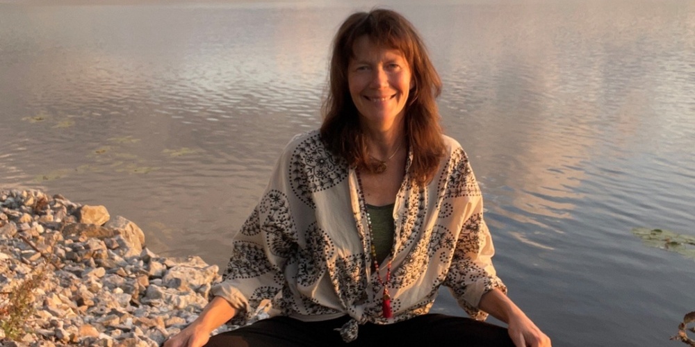 Kvinna sitter i meditationsställning på en brygga vid en sjö i solnedgången.