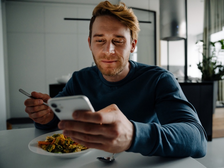 Man sitter vid ett bord, äter mat och tittar på sin mobiltelefon i ett modernt hem.