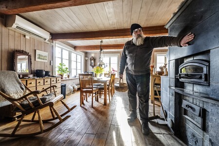 En man med skägg och mössa står vid en svart kamin i ett ljust, rustikt vardagsrum med trämöbler och trägolv.