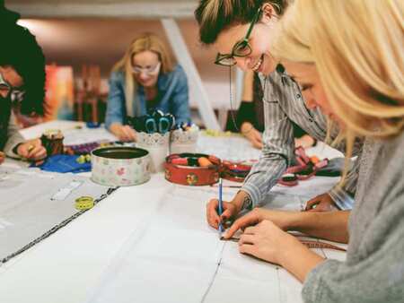 Två kvinnor designar ett mönster för att sy plagg. Två kvinnor arbetar i bakgrunden.