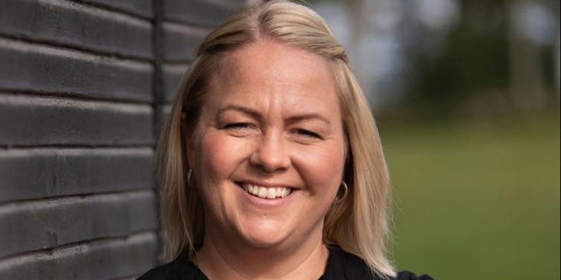 Leende kvinna med blont hår står med armarna i kors mot en svart trävägg utomhus.