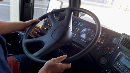 En person håller i ratten på en lastbil eller buss, med instrumentpanel och vägmiljö synlig utanför fönstret.