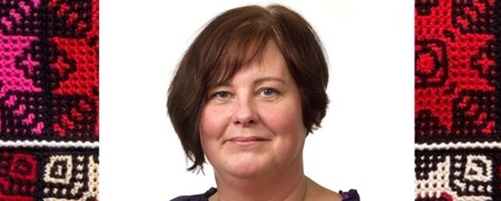 Kvinna med kort brunt hår och lila blus framför vit bakgrund med mönstrade textilier i rött och svart på sidorna.