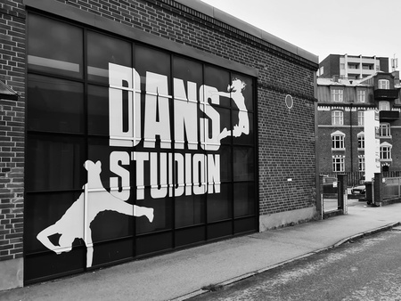 Svartvit bild på en dansstudio med stora fönster och texten 'DANS STUDIO' samt siluetter av dansare. Gata och äldre byggnader syns.