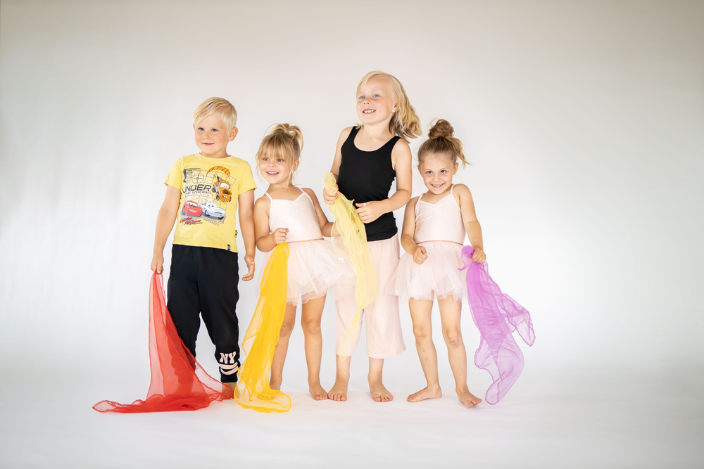 Fyra glada barn står tillsammans, tre i balettkläder och ett i t-shirt och byxor, med färgglada sjalar i handen.