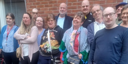 En grupp människor står och sitter utomhus framför en tegelvägg, några med färgglada kläder.