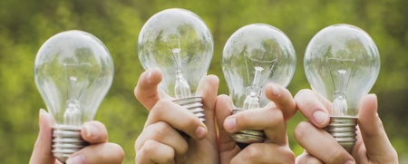 Fyra händer håller upp varsin glödlampa utomhus med grönskande bakgrund.